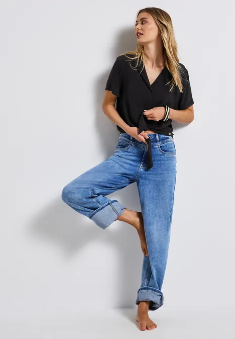 Frau in schwarzer Bluse und blauer Jeans steht barfuß an einer Wand.